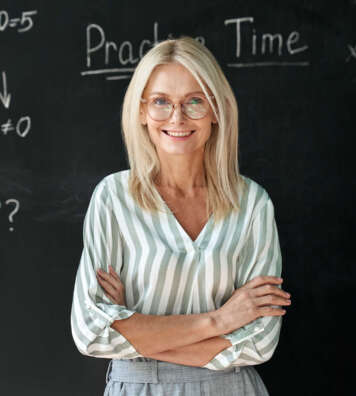Happy smiling middle aged woman mature high school education math teacher or college professor wearing glasses standing at blackboard background. Confident female professional educator portrait.