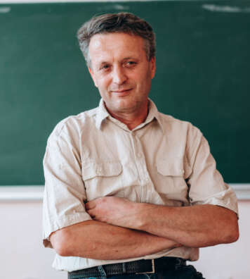 Happy teacher sitting folded a hands next a blackboard.- Image