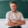 Happy teacher sitting folded a hands next a blackboard.- Image
