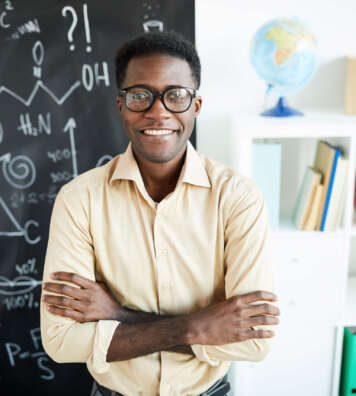 Young successful African-american teacher with arms crossed on chest standing by blackboard in classroom