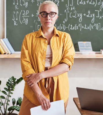 Portrait of confident teacher in eyeglasses standing in the classroom with blackboard in the background